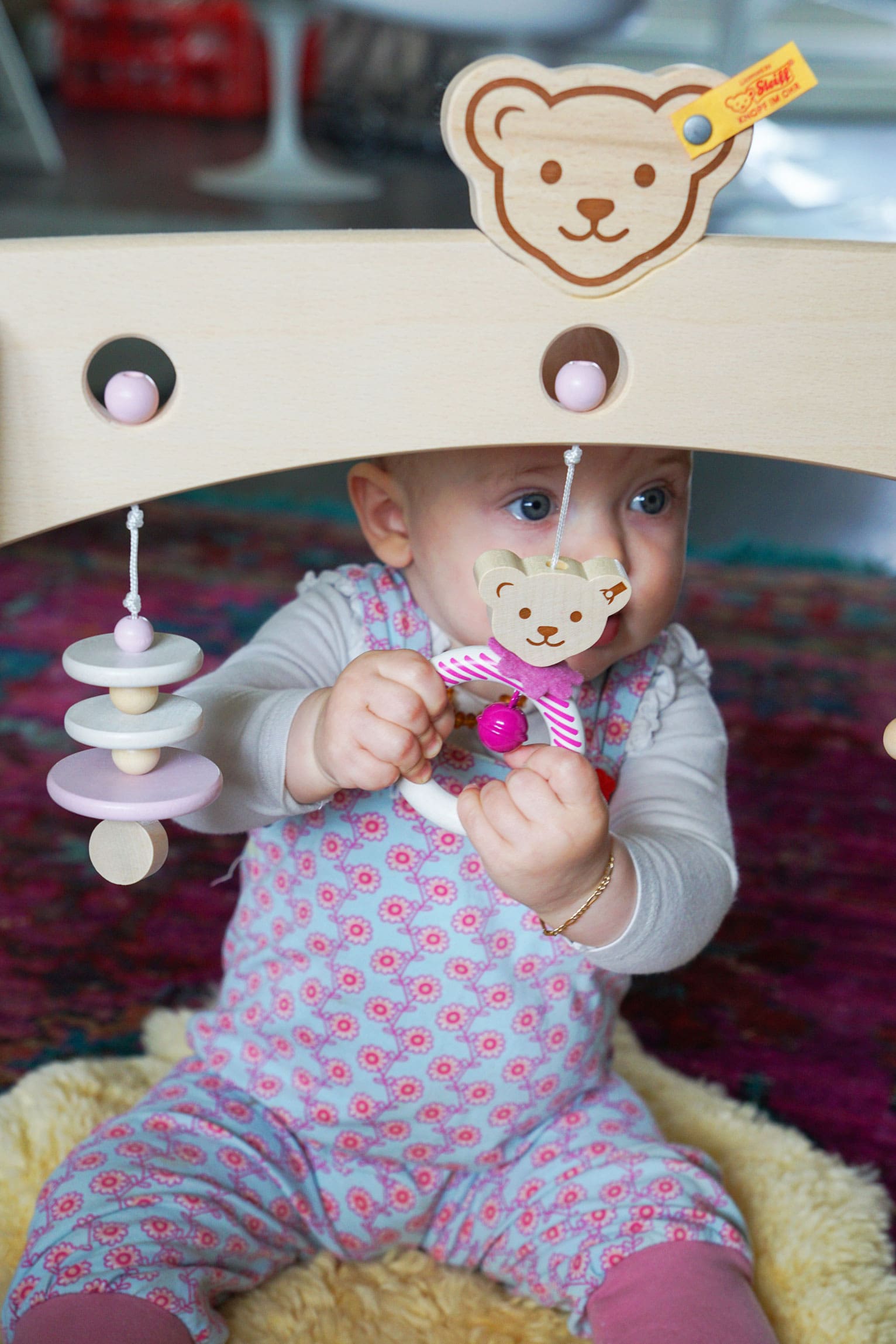 Baby sitzt auf einer Decke und spielt mit einem Steiff-Holzspielbogen; Blick konzentriert auf ein hängendes Bären-Spielzeug gerichtet.