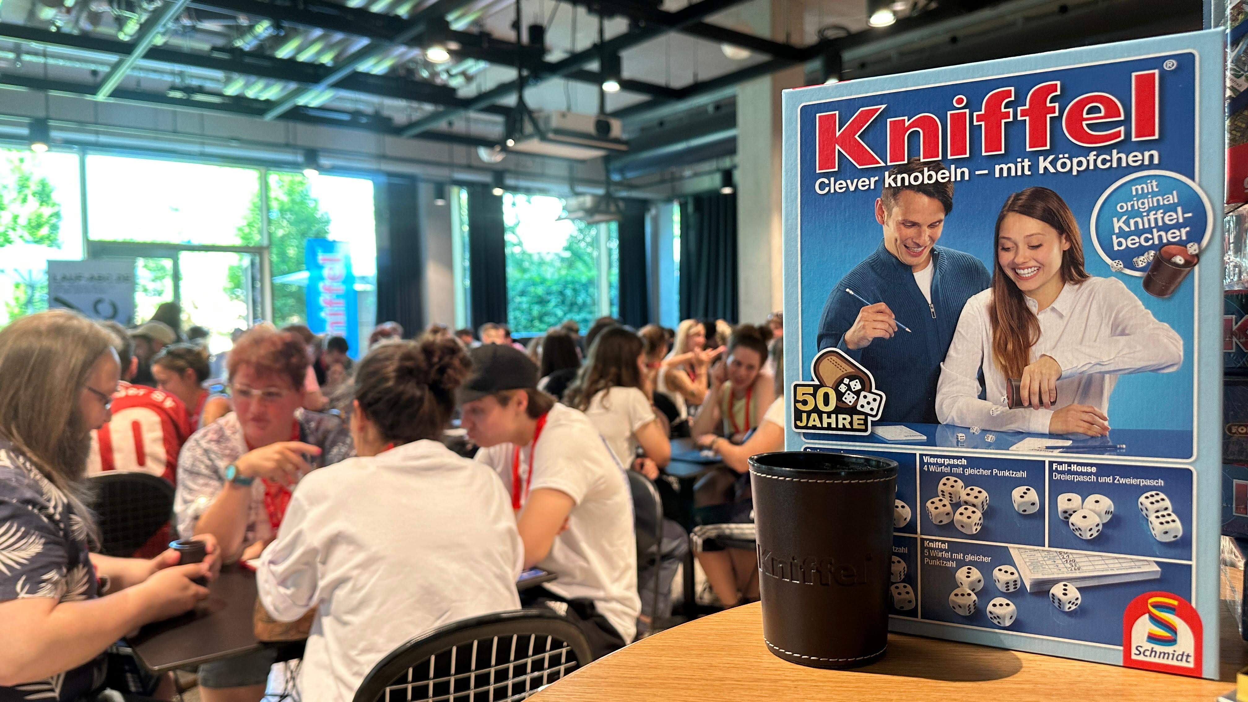 Kniffel-Spielzubehör: Würfelbecher, fünf Würfel, drei blaue Stifte und ein Spielblock mit Punktefeldern auf weißem Hintergrund.