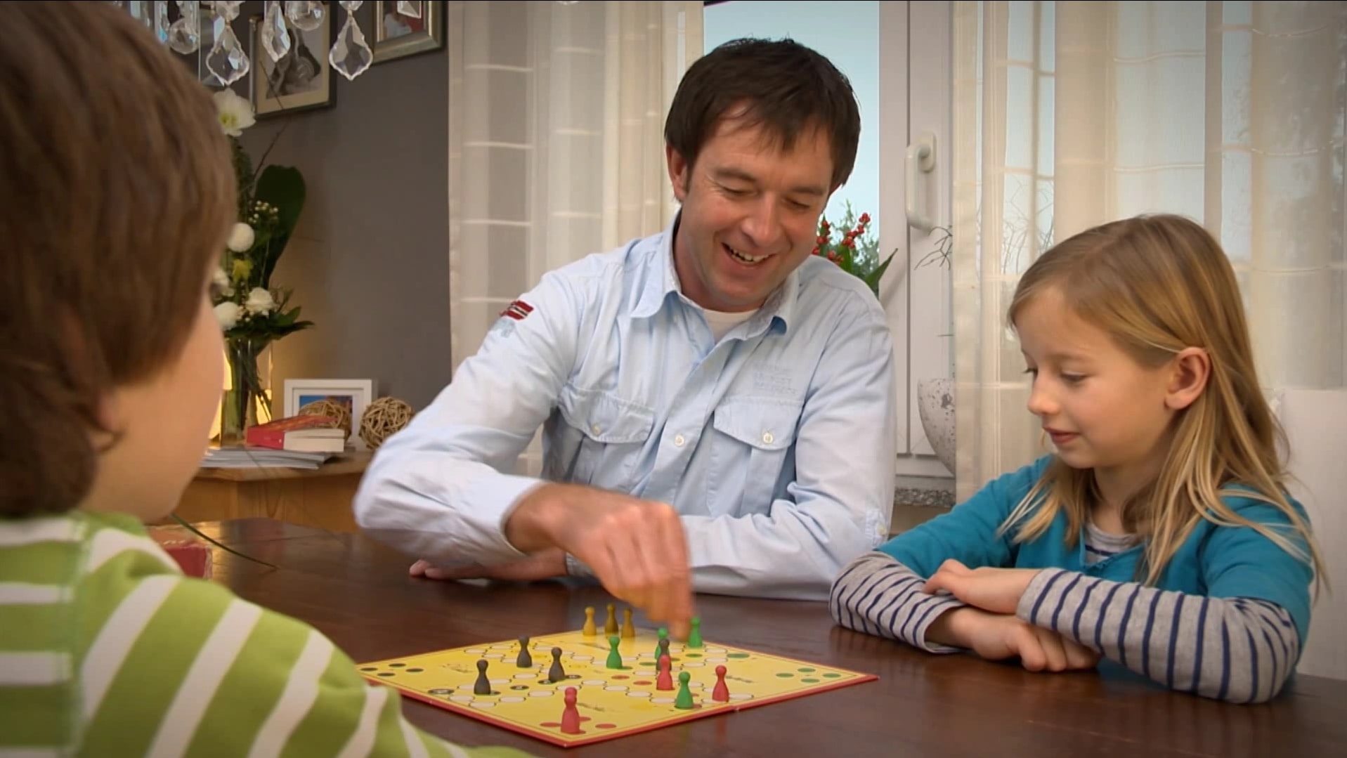 Ein Mann und zwei Kinder spielen gemeinsam ein Brettspiel an einem Holztisch in einem gemütlich eingerichteten Wohnzimmer.