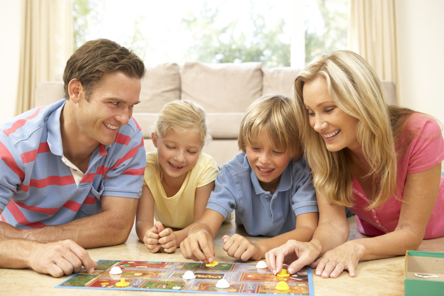 Eine lachende Familie liegt auf dem Boden und spielt gemeinsam ein Brettspiel mit bunten Spielfiguren.