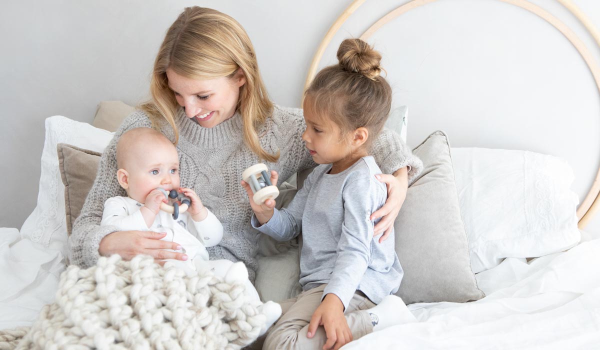 Mutter sitzt mit Baby und Kleinkind auf dem Bett, alle lächeln, das Baby hält ein Spielzeug, das Kind zeigt ein weiteres.