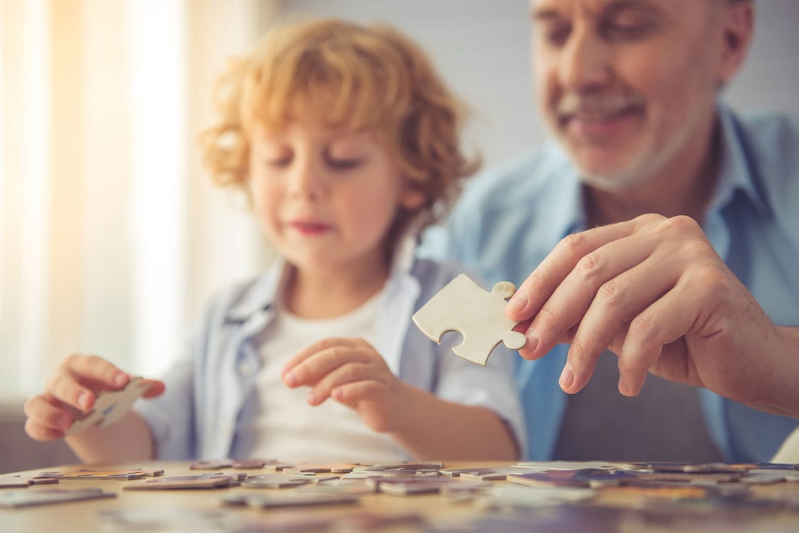 Kind und älterer Mann sitzen gemeinsam am Tisch und puzzeln, beide lächeln und halten Puzzleteile in der Hand.
