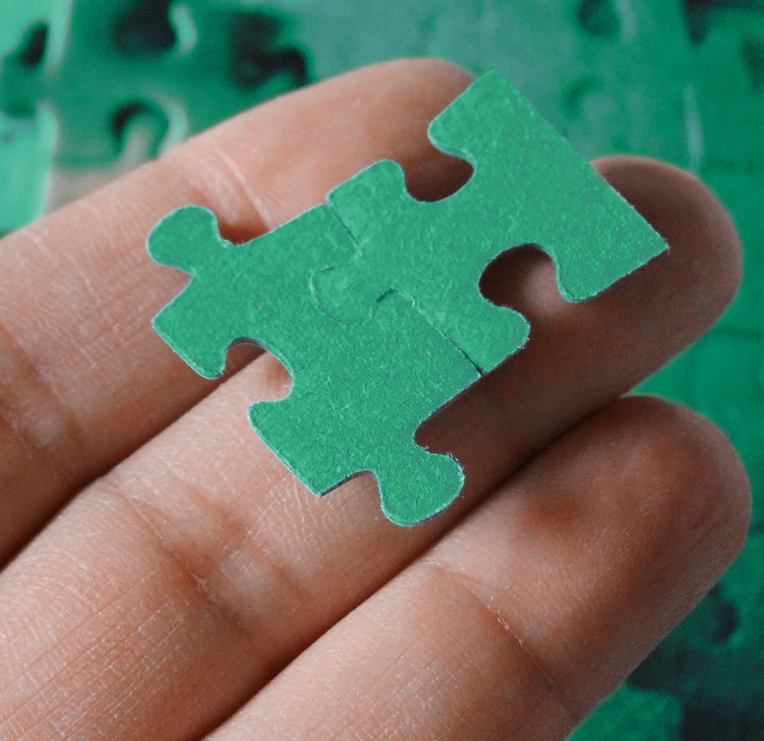 Nahaufnahme einer Hand, die zwei grüne Puzzleteile hält, die nahtlos ineinanderpassen; verschwommener Puzzleteppich im Hintergrund.